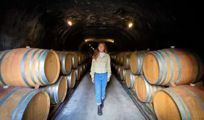 A woman explores a dimly lit wine cellar tunnel, walking between stacked barrels on a luxurious gourmet wine tasting tour experience.