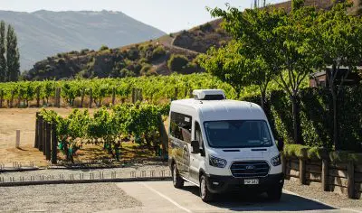 White van parked by vineyard under sunny skies, surrounded by lush green vines and rolling hills, ready for scenic wine tour.