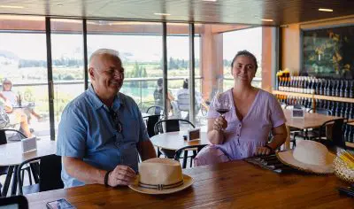 Couple enjoys premium Boutique Wine Tour, smiling at rustic wooden table indoors as woman savours a glass of red wine, exuding joy.