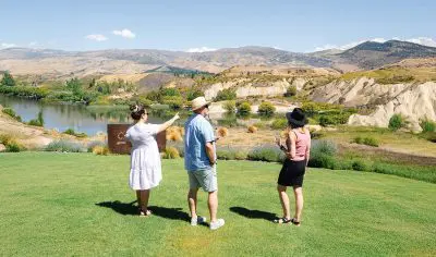 Three people enjoy a boutique wine tour, standing on lush grass and admiring a scenic lake and rolling hills under a clear blue sky.