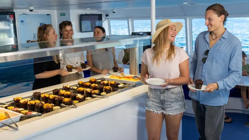 Guests on a Purely Whitehaven Day Tour select fresh pastries and muffins from a gourmet buffet, smiling and socialising together.