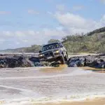 Black SUV navigates rugged terrain on a 3 Day Kgari Adventure near Rainbow Beach, with ocean waves and lush trees surrounding the vehicle.