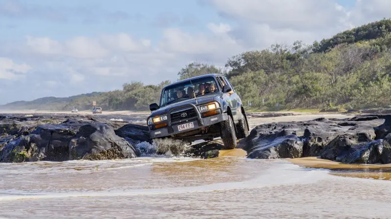 Black 4WD navigating rugged coastal rocks on a 3-Day K’gari adventure tour with dormitory accommodation, Rainbow Beach, Australia.