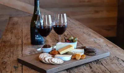 Elegant wooden table arranged for an upmarket wine tasting: two glasses, assorted cheeses, biscuits, dried fruit, olives, and chutney.