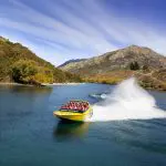 Experience the thrill as a yellow jet boat filled with excited passengers speeds across a vibrant blue river, spray erupting, lush hills and green trees behind.