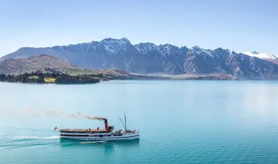 A historic steamship glides on a serene blue lake, offering a top-rated Classic Wine Tour with majestic mountains in the background.