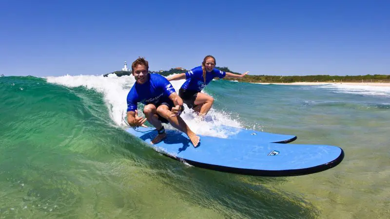 Children in matching blue shirts ride gentle, clear waves at Byron Bay's exclusive group surfing lesson by a sunlit sandy beach.