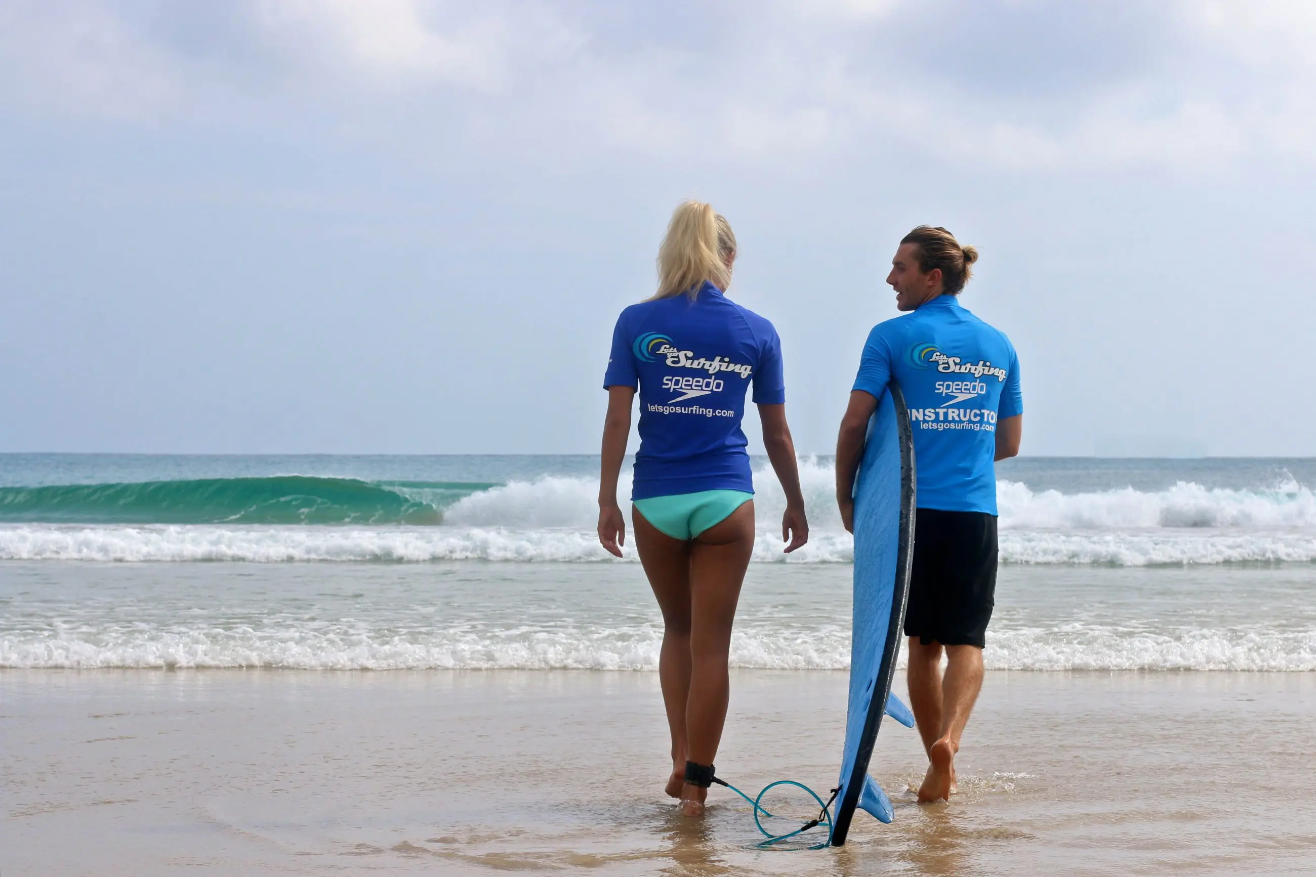 Two surfers in blue shirts on Byron Bay beach prepare for a 1.5-hour private surf lesson with Let's Go Surfing, watching the waves.