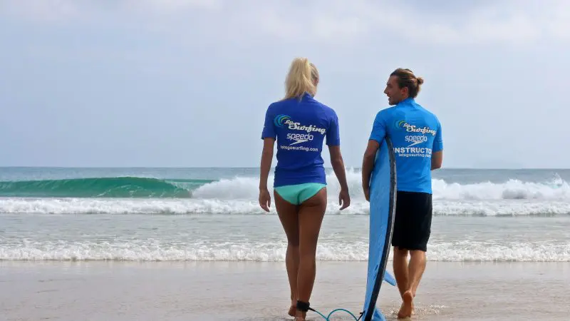 Two surfers in blue shirts on Byron Bay beach prepare for a 1.5-hour private surf lesson with Let's Go Surfing, watching the waves.
