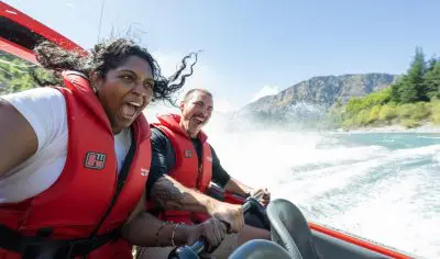 Two people in red life jackets enjoy a thrilling Shotover Jet ride, splashing water with stunning Queenstown mountains in the background.