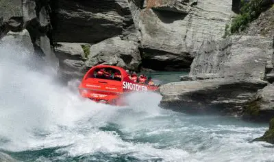 Experience the thrill as a red Shotover Jet surges through Queenstown’s rocky river rapids, helmeted riders gripping tightly on board.
