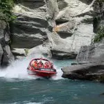 A bright red Shotover Jet boat races through a scenic rocky canyon over crystal-clear water, splashing mist as excited riders hold on.