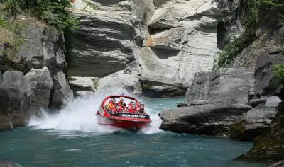 A bright red Shotover Jet boat races through a scenic rocky canyon over crystal-clear water, splashing mist as excited riders hold on.