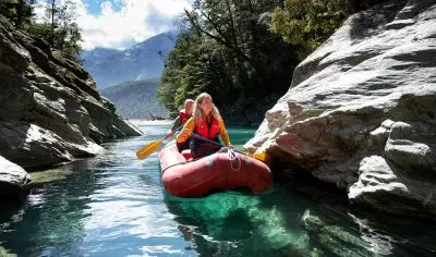 Two adventurers paddle a bright red Funyak down New Zealand’s crystal-clear Dart River, framed by dramatic rocky cliffs and lush forest.