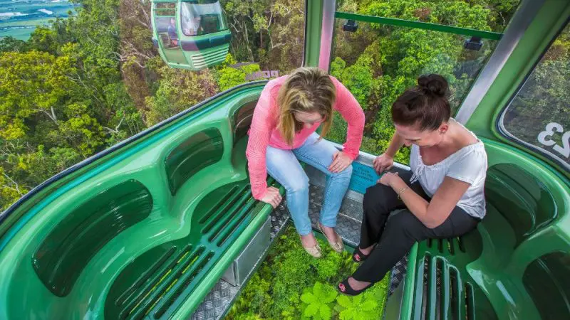 Two women gaze at lush rainforest vistas through a glass-floored cable car on the Express Kuranda Later Departure, experiencing scenic views.