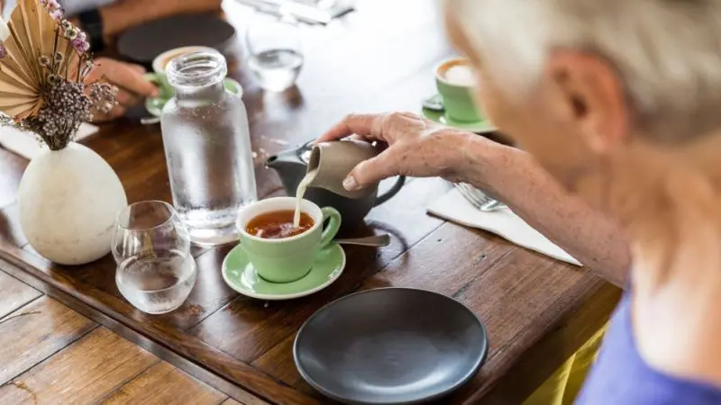 Pensioner in Cairns pours milk into tea at a rustic wooden table, surrounded by teacups, water glasses, and dried floral décor.