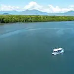 Experience a scenic Cairns City Sights River Cruise as a small white boat glides along a wide river, framed by lush greenery and mountains.