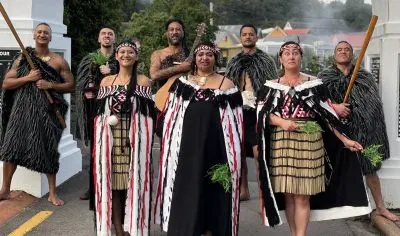 Māori group in traditional attire stands at Whakarewarewa, Rotorua, outdoors, displaying paddles and native greenery for a photo.