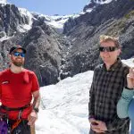 Three adventurers in outdoor gear smile on snowy ground during a Glacier Encounter, with sunlit rocky mountains in the background.