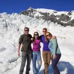 Four friends smile on a snowy Glacier Encounter, stunning mountains and vibrant blue sky behind them for the perfect outdoor adventure.