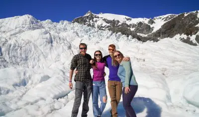 Four friends smile on a snowy Glacier Encounter, stunning mountains and vibrant blue sky behind them for the perfect outdoor adventure.