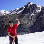 Man in red shirt stands on snowy glacier, smiling amid majestic mountain peaks and vibrant blue sky, enjoying an epic adventure.