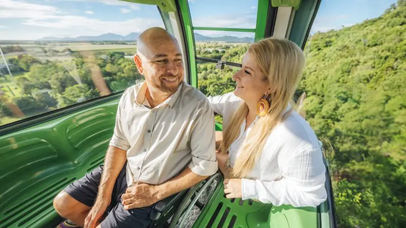 Smiling man and woman ride a green cable car on the 3 Day Daintree Cape Tribulation Port Douglas Comfort Tour, enjoying scenic views.