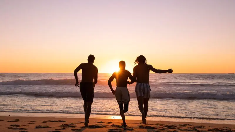Three travellers dash towards the ocean at vibrant sunset, embodying adventure on a 3 Day Daintree Cape Tribulation Comfort Tour.