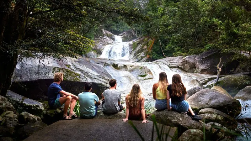 Six travellers unwind on rocky terrain beside a scenic waterfall during the 3 Day Daintree Cape Tribulation Port Douglas Comfort Tour.