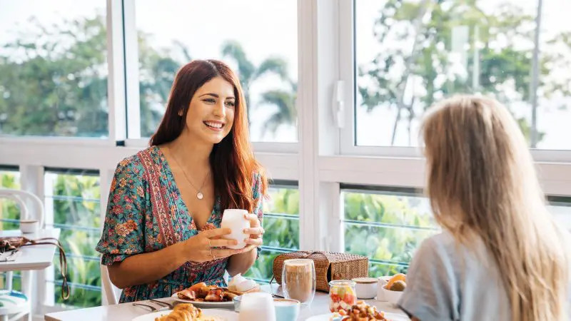 Two women have breakfast in a sunlit café, discussing their 3 Day Daintree, Cape Tribulation & Port Douglas Comfort Tour adventure.