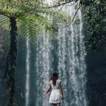 A woman in a flowing white dress stands beside a cascading waterfall, evoking the 16 Day Best Of East Coast Australia Comfort Tour experience.