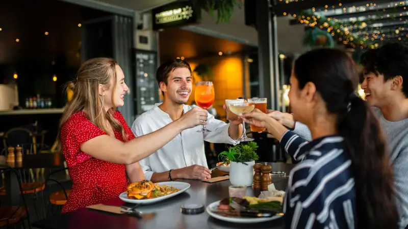 Four friends raise glasses in a lively restaurant, celebrating their 16-day Best of East Coast Australia Comfort Tour from Sydney.