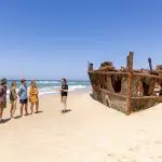 Travellers relax on a pristine Fraser Island beach by a historic shipwreck during the 8 Day Sydney to Brisbane Comfort Tour adventure.