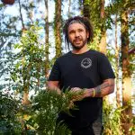 Smiling man in black shirt surrounded by lush green plants on the 16 Day Best Of East Coast Australia Comfort Tour, enjoying nature.