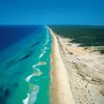 Aerial view of a pristine sandy beach on the 8 Day Sydney to Brisbane with Fraser Island Comfort Tour, turquoise waves, grassy shore.