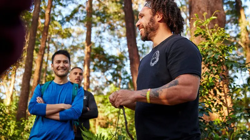 Three men laughing and chatting in a sun-dappled forest on a 14 Day Sydney to Cairns Comfort Tour, surrounded by vibrant greenery.