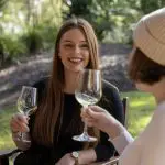 Two women smiling and toasting glasses of wine in a lush park during their 16 Day East Coast Australia Comfort Tour from Sydney.