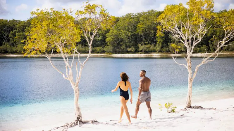 A man and woman walk along a scenic sandy lake beach, capturing the essence of a 14-Day Sydney to Cairns Comfort Tour adventure.