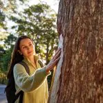A woman in a green jumper touches a tree trunk in a sunlit forest during her 8 Day Sydney to Brisbane Fraser Island adventure tour.