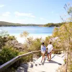 Two travellers hike sandy steps by lush trees, overlooking a blue lake and rolling hills on the 8-Day Sydney to Brisbane Fraser Island Tour.