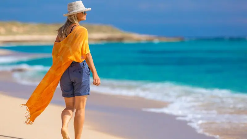 Barefoot woman in sun hat and vibrant orange shawl walks along pristine beach—discover Coastal Wonders on 8 Day Perth to Exmouth Tour.