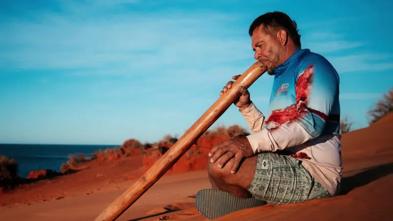 Man playing didgeridoo on red sand beach, experiencing scenic coastal views on the 8 Day Perth to Exmouth Comfort Tour.