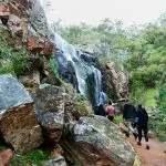 Three hikers trek a rugged Grampians trail towards a scenic waterfall, surrounded by lush greenery on an eco wilderness escape day tour.