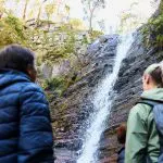Two people in jackets admire towering cliffs by a rocky waterfall during a 1 Day Grampians Eco Wilderness Escape, surrounded by nature.