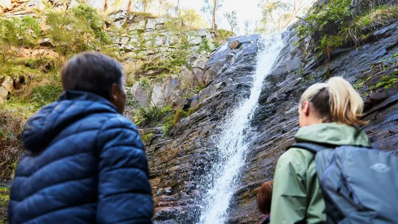 Two people in jackets admire towering cliffs by a rocky waterfall during a 1 Day Grampians Eco Wilderness Escape, surrounded by nature.