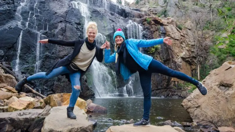 Two women enjoying a playful moment on rocks by a stunning Grampians waterfall during their 1 Day Eco Wilderness Escape adventure.