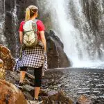 A female adventurer with a rucksack stands on rocky terrain, gazing at a stunning Grampians waterfall during her eco wilderness escape.