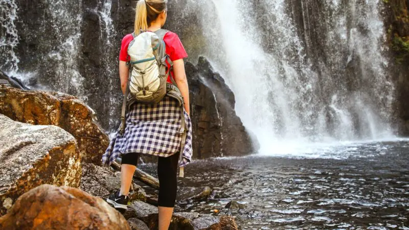A female adventurer with a rucksack stands on rocky terrain, gazing at a stunning Grampians waterfall during her eco wilderness escape.