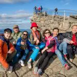 A group of seven happy people on rocky terrain enjoying a 1 Day Grampians Eco Wilderness Escape with stunning scenic views behind them.