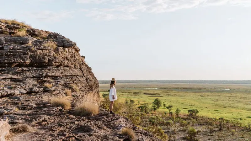 Discover the thrill of a 1 Day Kakadu Wilderness Escape: person in white atop rocky cliff overlooking lush, scenic grassy plains.